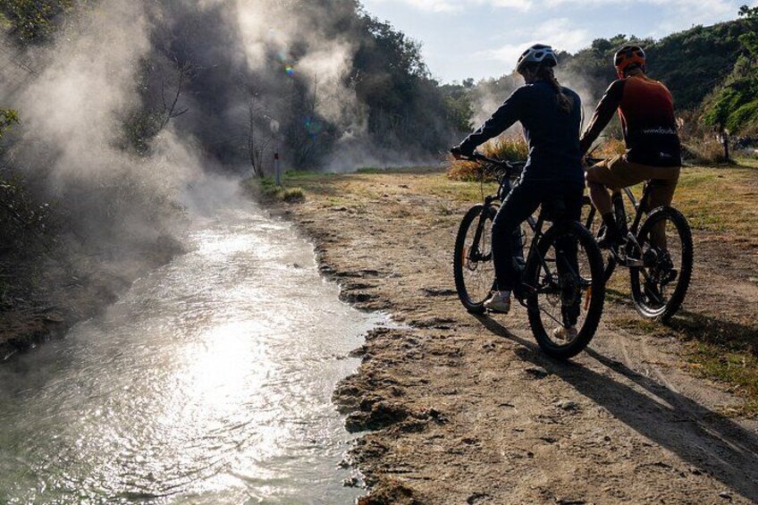Two cyclists on a dirt path beside a river with steam rising from the water. 2