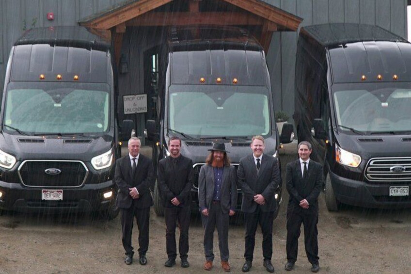 Five men in suits standing in front of three black trucks. 5