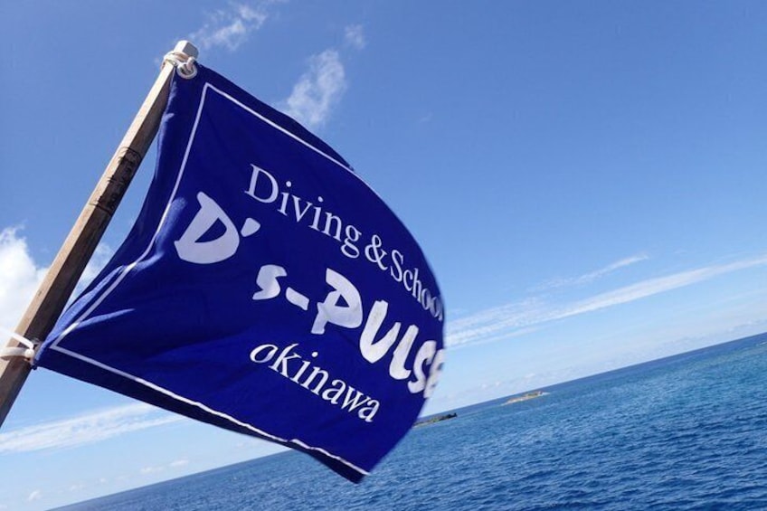 A flag with 'Diving & School' and 'D-S-PULS Okinawa' written on it, set against a clear blue sky and ocean. 3