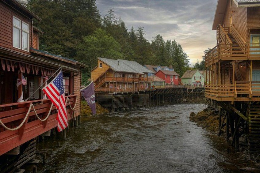 Una casa flotante de madera amarrada en un río, rodeada de otras casas sobre pilotes, con una bandera estadounidense y una bandera morada. 2