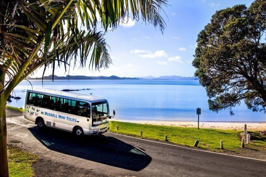 A white bus with 'Russell Mini Tours' on the side parked on a road with a beach and ocean in the background. 5