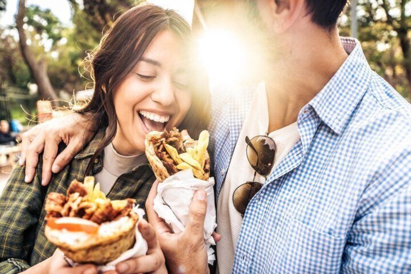 Two people holding food items, possibly sandwiches or wraps, outdoors with trees in the background. 3