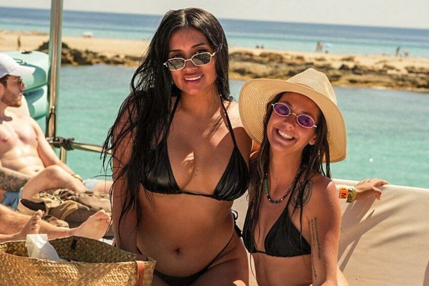 Two people in bikinis on a boat at a beach with clear blue water and sandy shore. 2