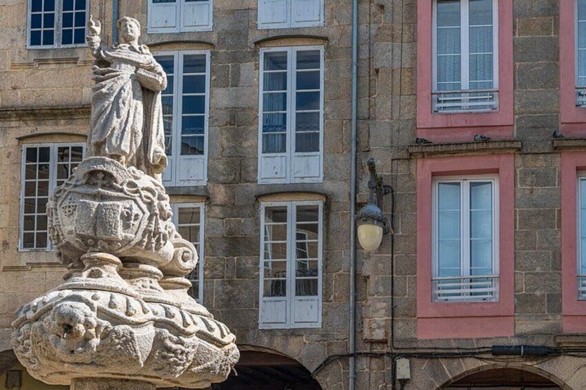 A stone statue with a figure holding a staff, set against a building with multiple windows and a street lamp. 5
