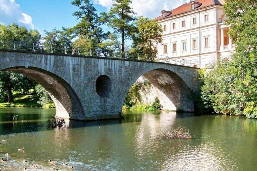 A stone bridge over a river with ducks swimming below, surrounded by trees and a building with a red roof. 1