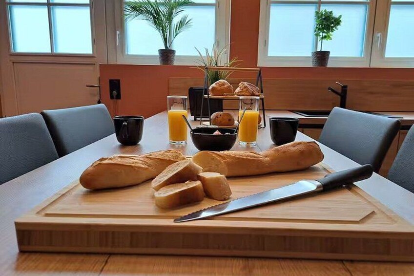 A wooden cutting board with bread, a knife, a glass of orange juice, and a bowl of fruit on a table. 4