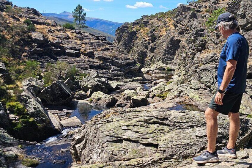 A rocky terrain with a stream, a person standing on a rock, and a mountain in the background. 2