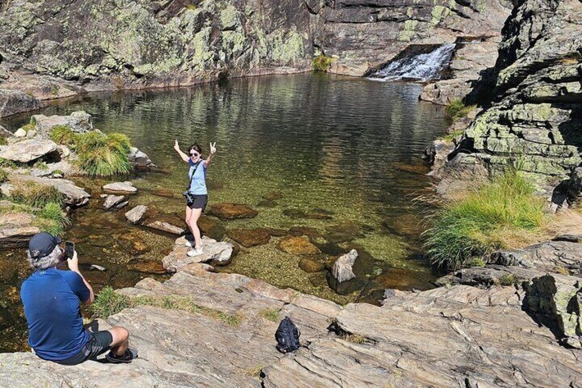 A person standing on rocks by a clear water body with a waterfall, another person taking a photo, and a backpack on the ground. 1