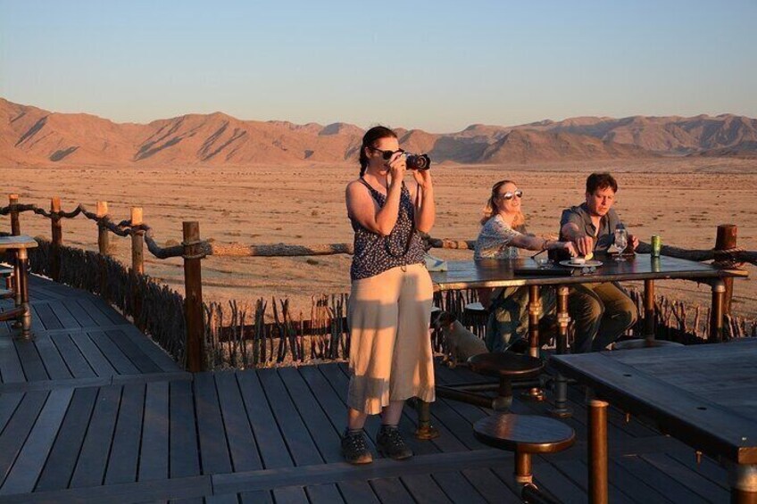 A wooden deck with tables and chairs, desert landscape in the background, and a person taking a photo. 3