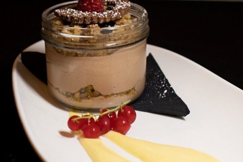 A dessert in a glass jar with a raspberry on top, accompanied by red grapes and a piece of yellow fruit on a white plate. 4