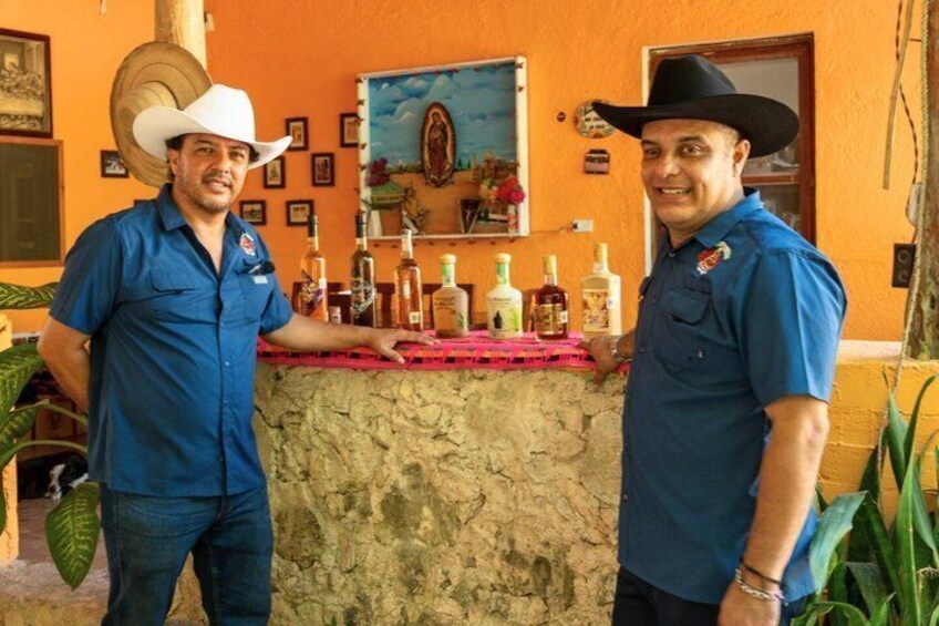 Two men in blue shirts and cowboy hats standing in front of a bar with bottles. 1