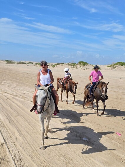 Three people riding horses on a sandy beach with dunes in the background. 3