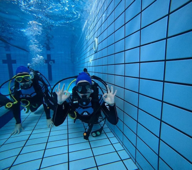 Two scuba divers in a tiled pool with blue tiles and bubbles visible. 1