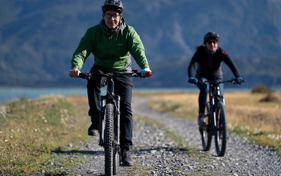 Two cyclists on a gravel path with a lake and mountains in the background. 3