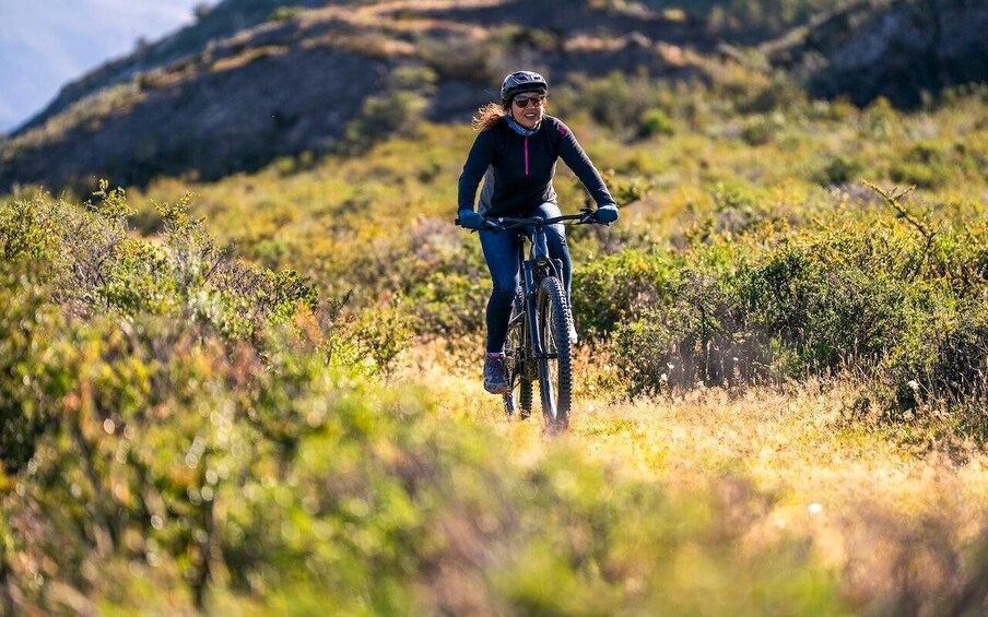 A person riding a mountain bike on a grassy trail with bushes and a rocky hill in the background. 2