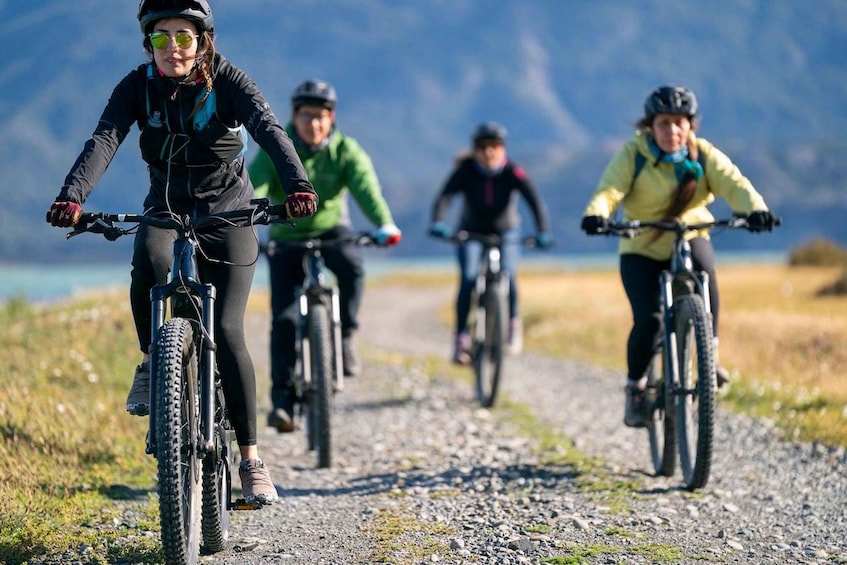 Four cyclists on a gravel path with a lake and mountains in the background. 1