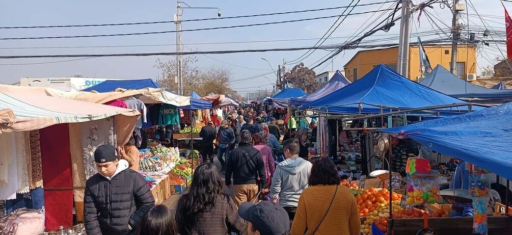 A crowded outdoor market with people shopping and vendors selling goods under blue and beige canopies. 1