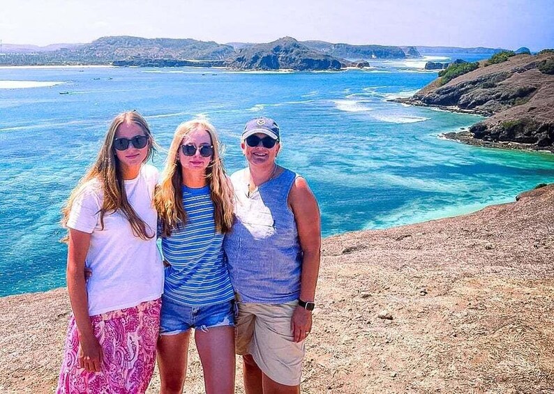 Three people standing on a rocky beach with a view of the ocean and distant hills. 2