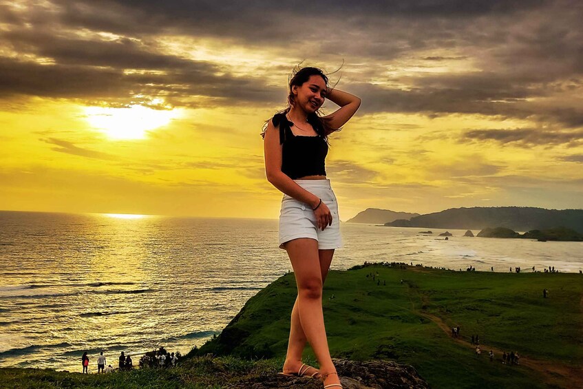 A person standing on a grassy hill overlooking a body of water with a sunset in the background. 1