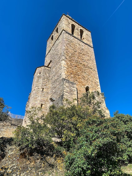 A tall stone tower with arched windows and a cross on top, surrounded by greenery. 3