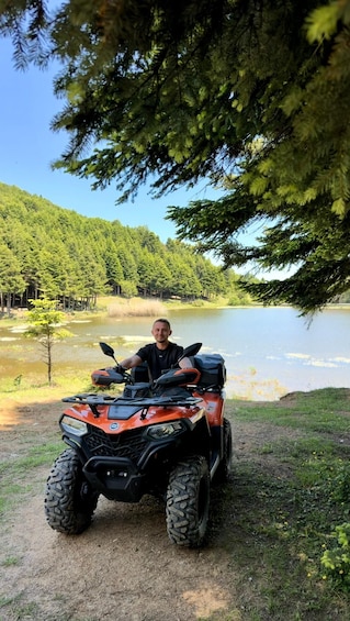 A person sitting on an orange ATV near a lake surrounded by trees. 4