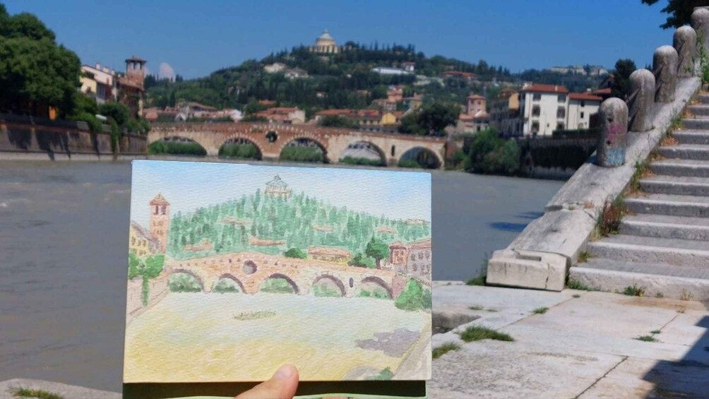 A hand holding a watercolor painting of a bridge over a river, with a cityscape in the background. 5