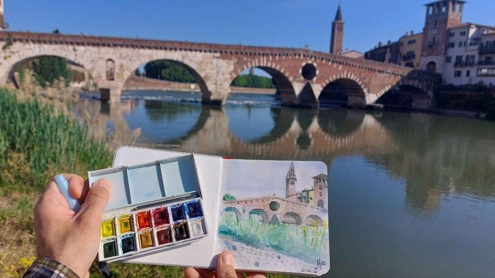 A hand holding a watercolor palette and a painting of a bridge over water. 1