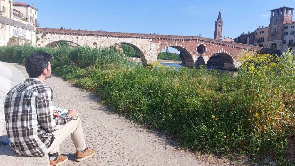 A man sitting on a path beside a river, overlooking a stone bridge with arches and a tower in the background. 2