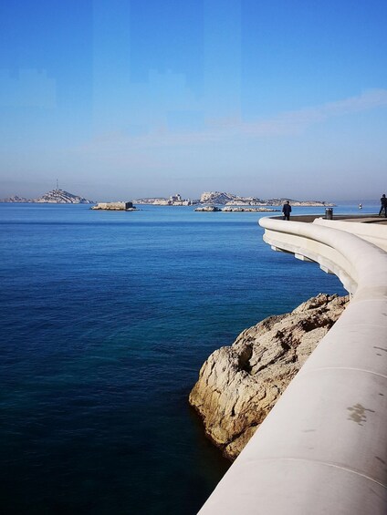 A coastal scene with a clear blue sky, a curved white structure, rocky outcrops, and distant islands. 2
