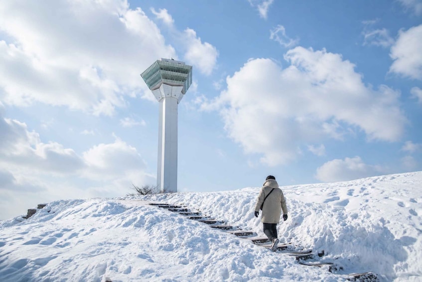 A person walking up a snowy hill towards a tall white tower with a glass top. 1