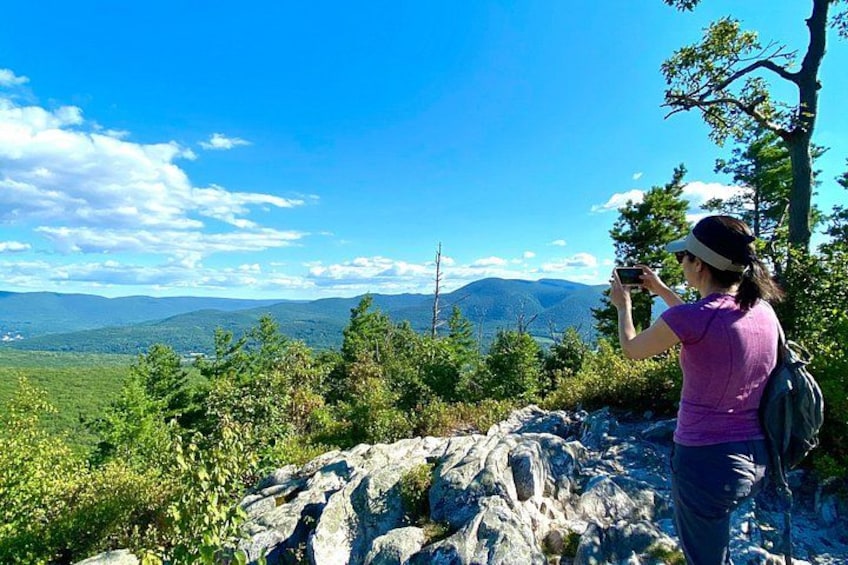 A person taking a photo with a smartphone, standing on a rocky outcrop with a view of mountains and a clear sky. 3