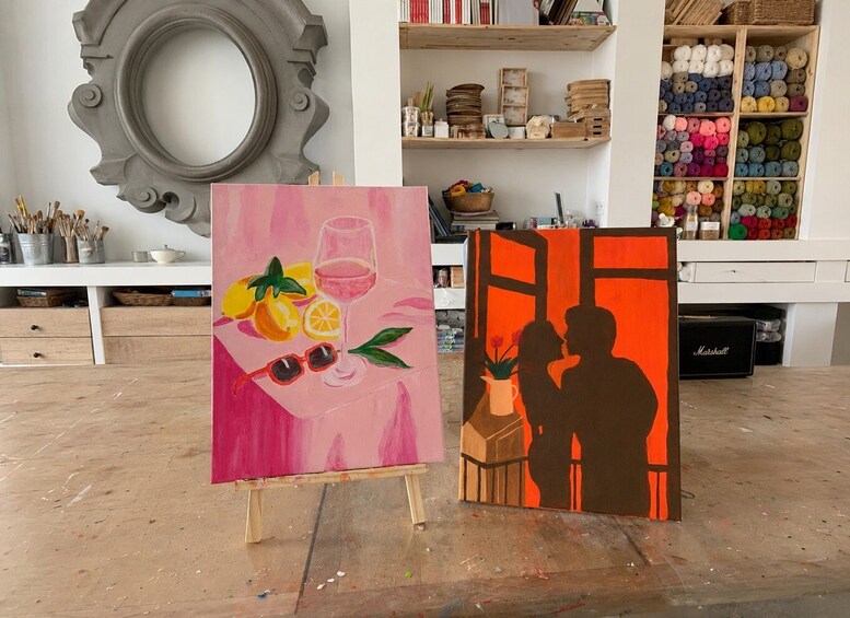 Two paintings on easels, one with a pink background and fruit, the other with a brown background and a couple. 3