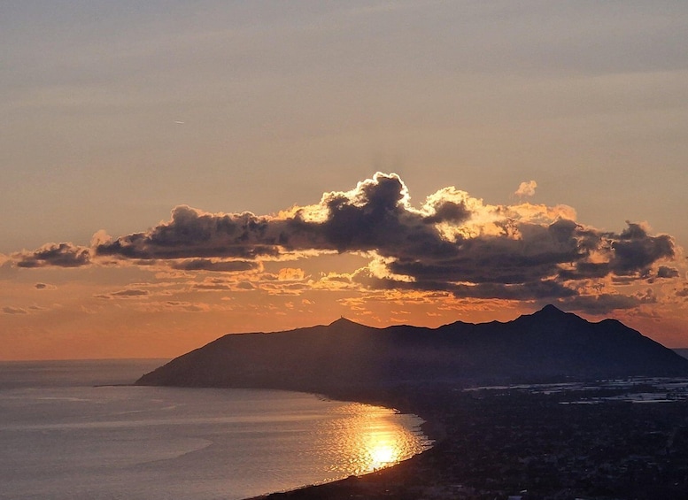 A sunset over a coastal landscape with mountains in the background and a calm sea reflecting the sunlight. 1