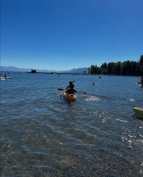 A kayaker paddling on a calm lake with clear water, surrounded by a forest and distant mountains. 4