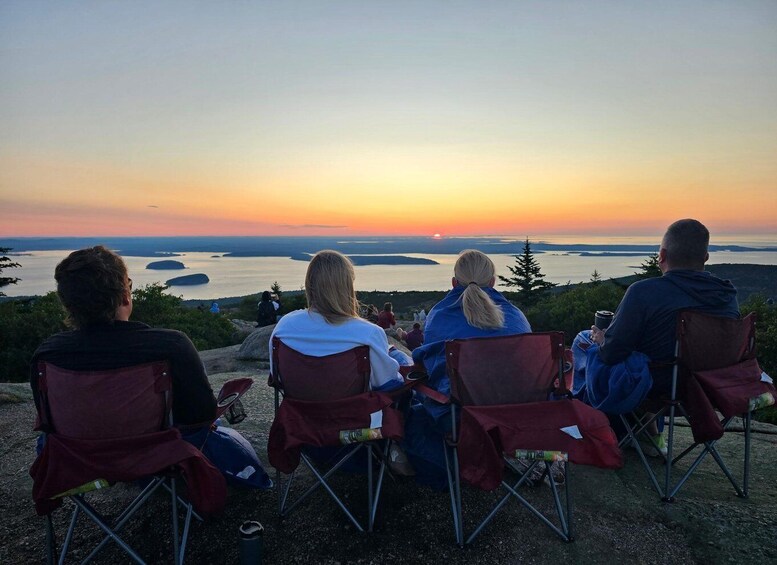 Three people sitting on camping chairs overlooking a sunset with a body of water and hills in the background. 5