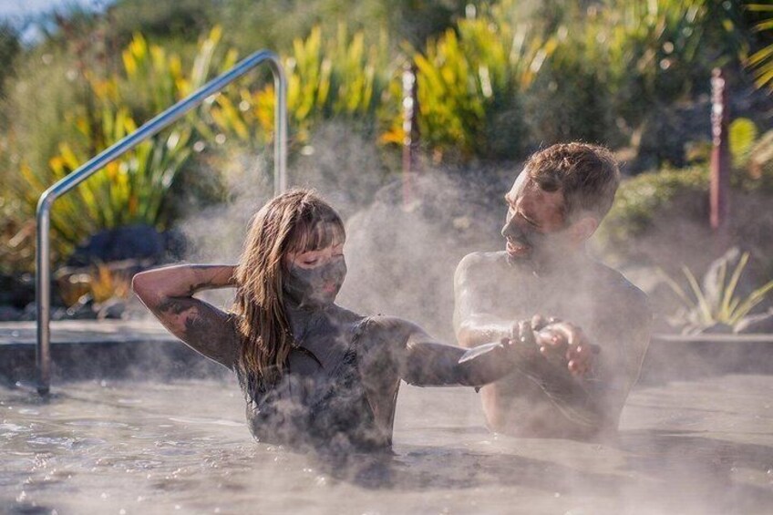 Two people in a hot spring with steam rising around them, surrounded by greenery and a metal railing. 1