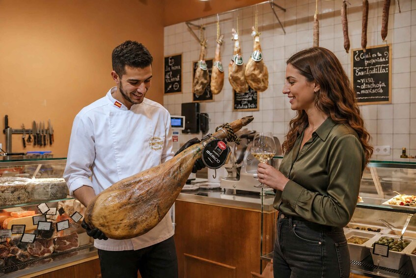 A person holding a large cured ham, a glass of wine, and another person in a restaurant setting. 5