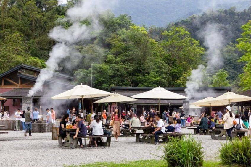 A group of people dining outdoors with steam rising from a cooking area, surrounded by trees and a mountainous backdrop. 1