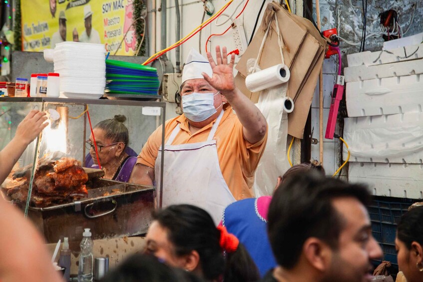 A street food vendor with a hand raised, surrounded by cooking equipment and a crowd. 3