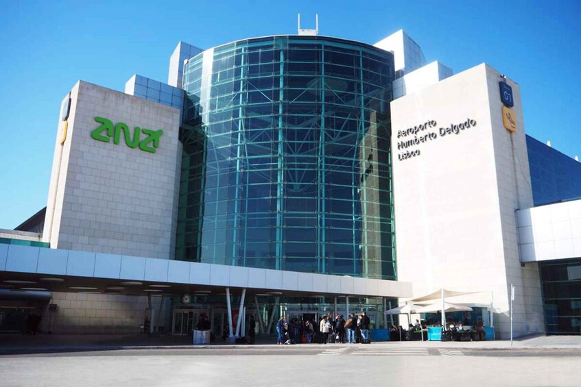 A modern building with a curved glass facade and signage for 'Aeroporto Humberto Delgado Lisbon'. 1