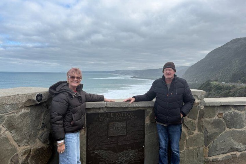 Two people standing by a stone wall with a plaque that reads 'Cape Point The Great Ocean Road.' 2