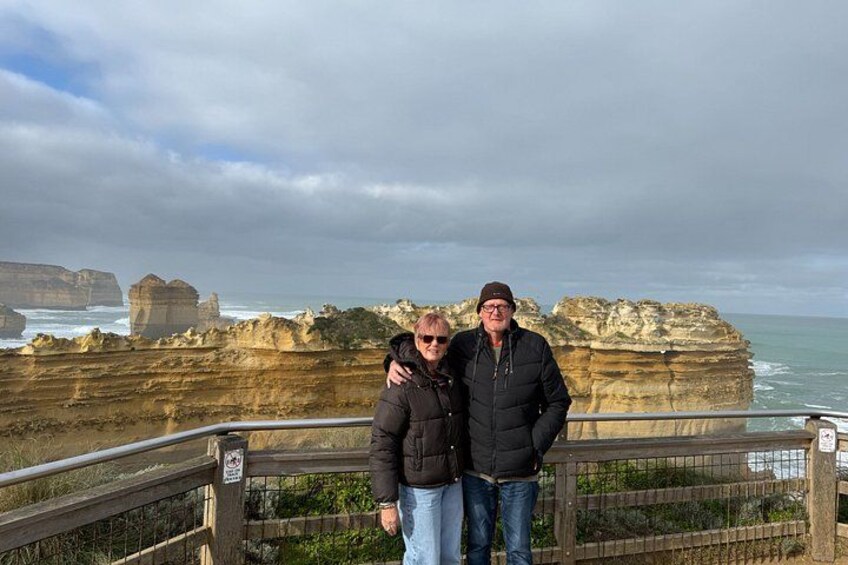 Two people standing on a wooden boardwalk with a coastal cliff landscape in the background. 5