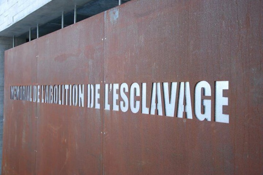 A large metal wall with the inscription 'Memorial de l'abolition de l'esclavage' in white letters. 2