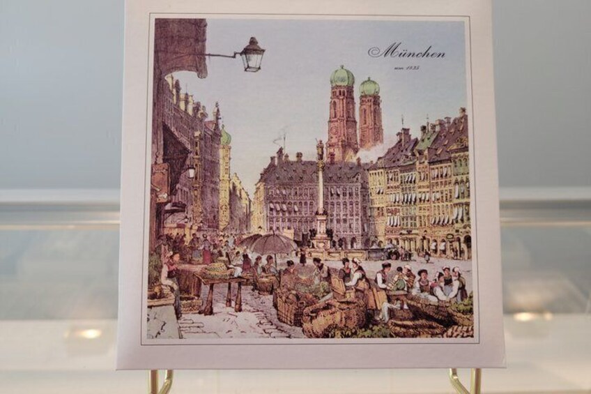 A framed illustration of Munich's historic center with buildings, a market scene, and the Frauenkirche. 4