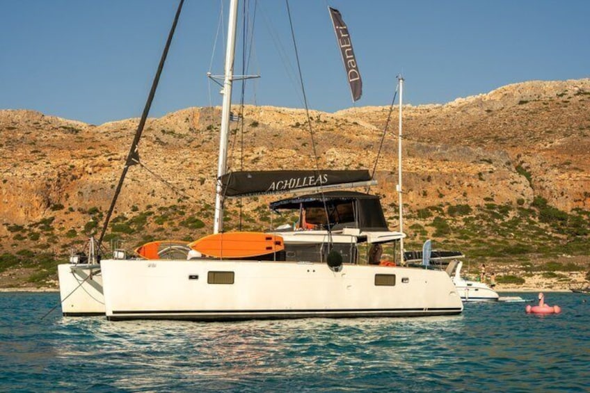 A white yacht with an orange deck, named 'ACHILLEAS,' anchored near rocky hills. 4