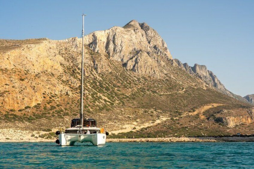 A white boat with a tall mast sailing on turquoise water, with a rocky mountain in the background. 1
