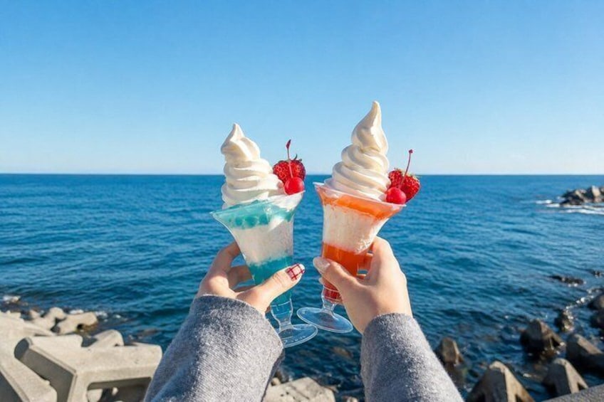 Two ice cream cones with toppings, held by hands against a seaside backdrop. 5