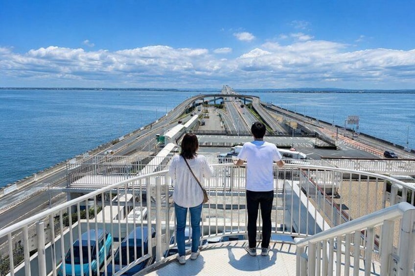 Two people standing on a bridge railing overlooking a coastal road with a clear sky. 4