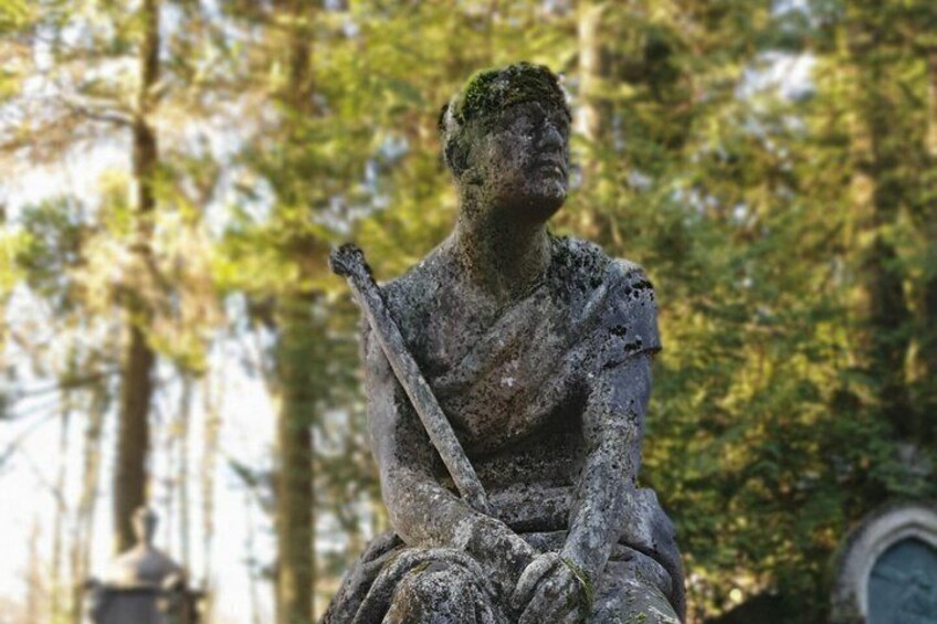A weathered statue of a seated figure holding a staff, surrounded by trees. 3