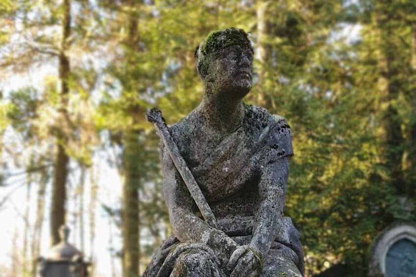 A weathered statue of a seated figure with a sword, surrounded by trees. 3